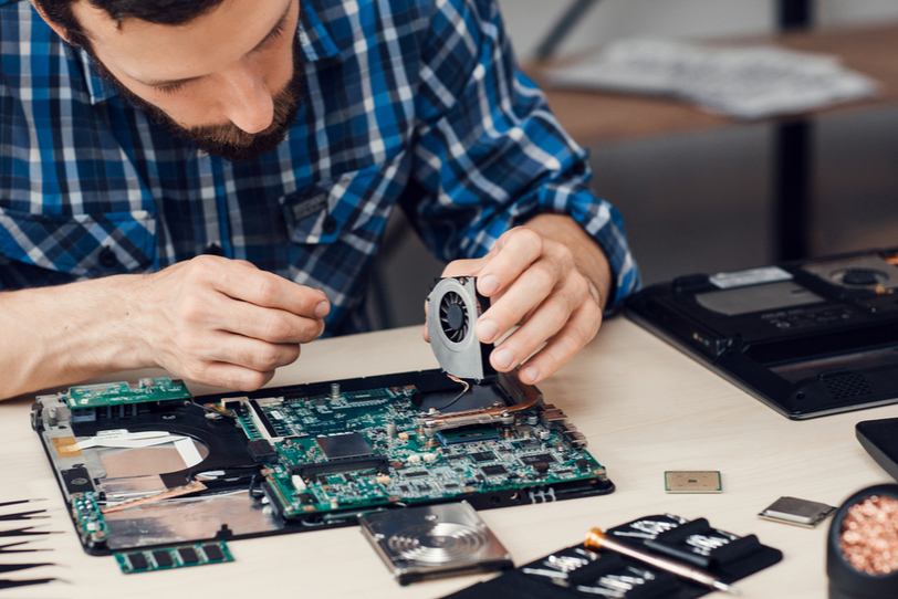 laptop repairng Technician repairing a laptop by carefully installing or removing a cooling fan from the motherboard, with various electronic tools and components spread out on the desk.